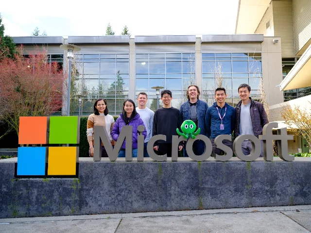 七名 Cove 团队成员站在 Microsoft 标识后方，前景放着 Cove 的绿色吉祥物
