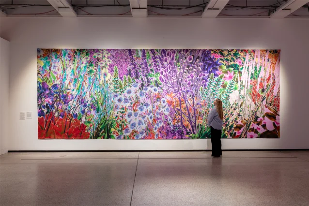 A large vividly coloured botanical tapestry showing dense flowers and plants across a museum wall with a visitor standing in front