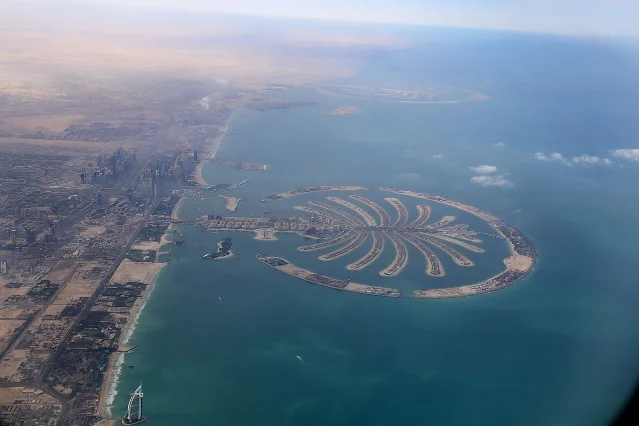 Aerial photograph showing the full Palm Jumeirah structure including trunk, 17 fronds, and crescent breakwater, with the Dubai skyline and Burj Al Arab visible to the south