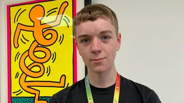 Young man in a black shirt with a lanyard standing in front of a vibrant artwork poster.