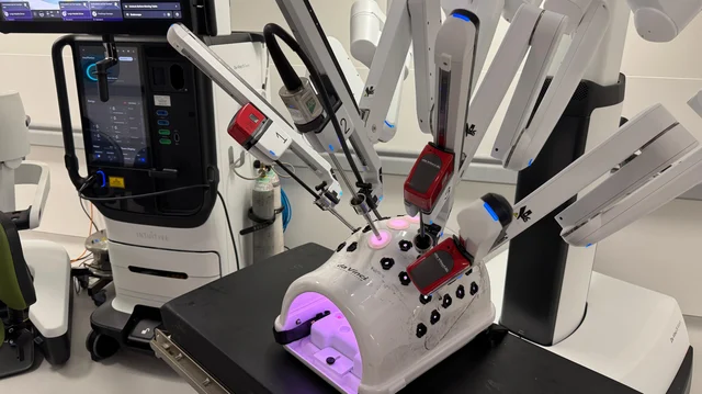 a white surgical robot with several jointed arms reaching down towards a training dome, beside a console with a screen