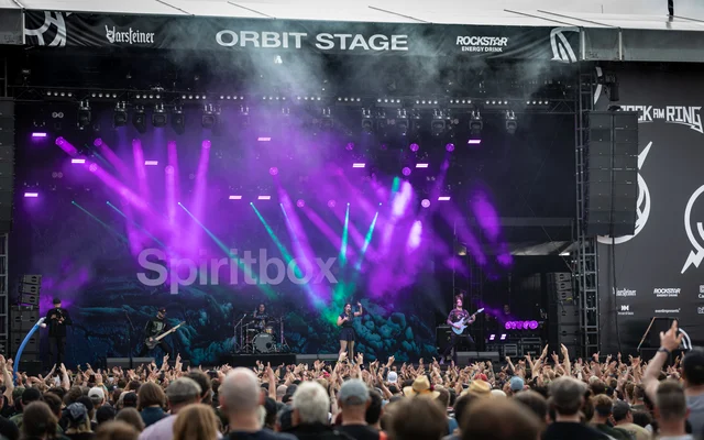 A band playing a large outdoor festival stage seen from above the crowd, with purple spotlights and the band's name on the backdrop