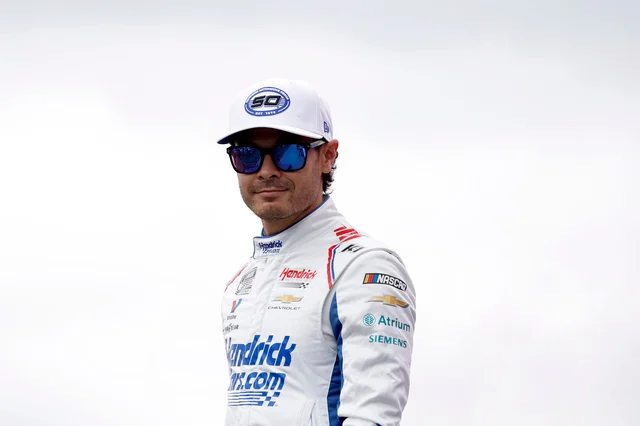 Kyle Larson in a white Hendrick Motorsports race suit and cap, wearing mirrored sunglasses
