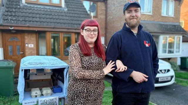 a woman and a man standing beside a small covered egg stall in a front garden