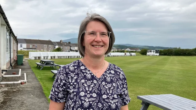 Smiling woman with short grey hair and glasses in a floral top standing in front of a village green and cricket pitch.