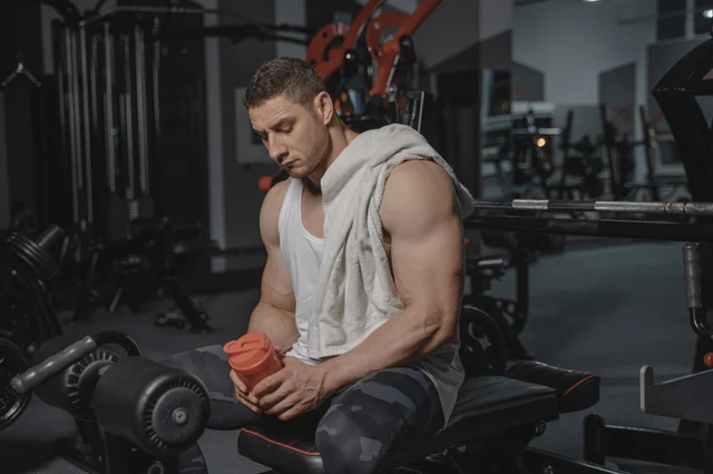 Man at gym sitting on bench holding a protein shaker after a workout, towel over shoulder