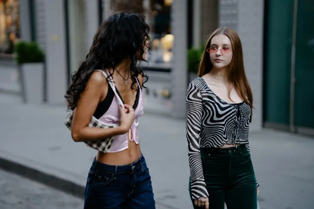 Two women in coordinated street looks, one in a sheer patterned mesh crop top and one in a silky satin crop with exposed bralette