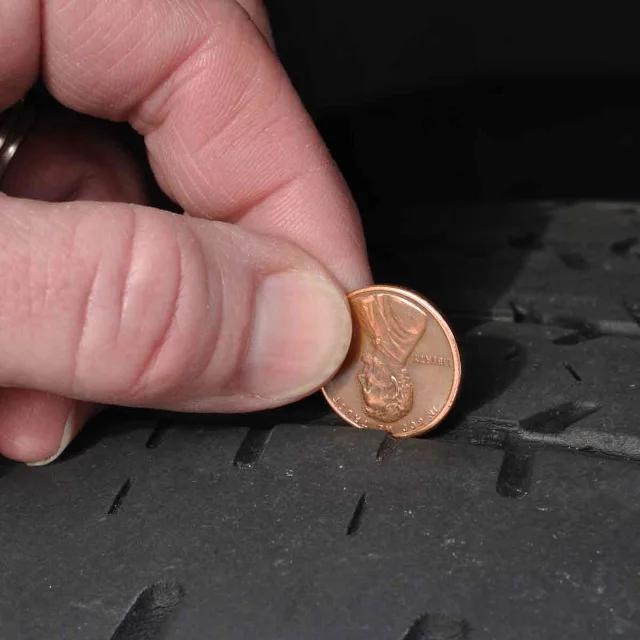A hand pressing a penny into a worn tire's tread groove, with the coin's head upside down