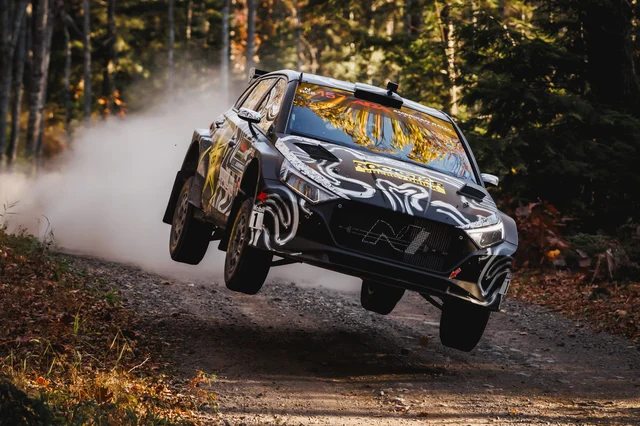 A rally car airborne over a gravel forest road, throwing up dust