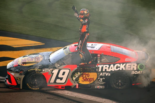 Chase Briscoe standing on the roof of his No. 19 Joe Gibbs Racing NASCAR Cup car with a raised fist, tires smoking