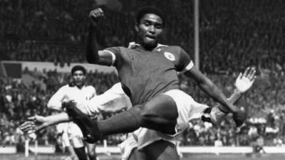 Eusébio in a dark Portugal shirt swings at the ball as players in white close in, with a packed stand behind them.
