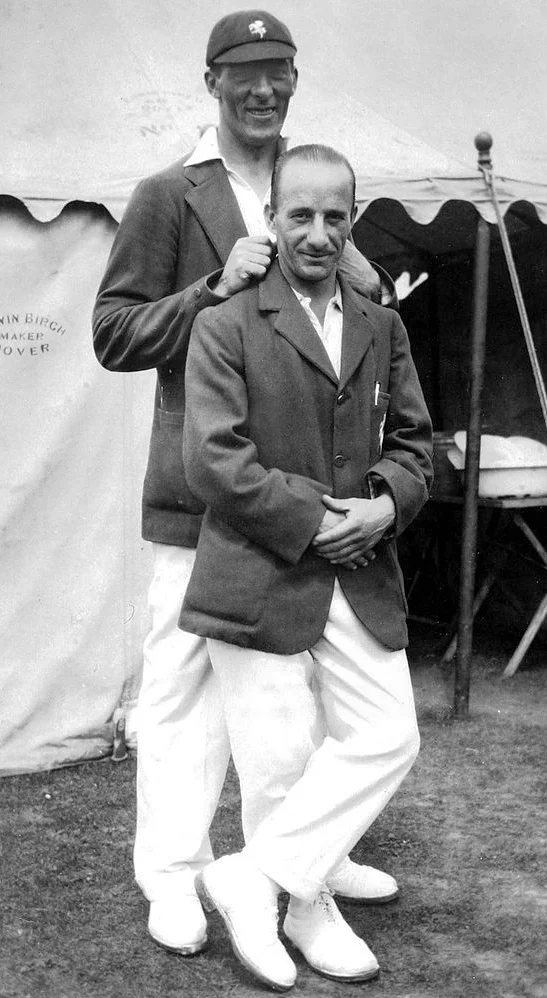 Two cricketers in whites and dark blazers standing outside a marquee, the shorter man in front with his hands clasped
