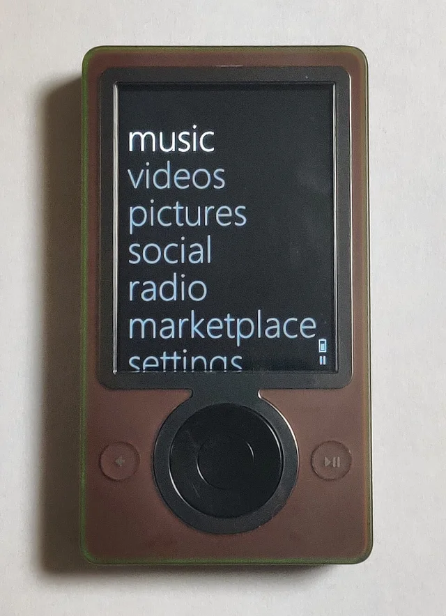 棕色外壳的 Microsoft Zune 30 正在显示主菜单界面,屏幕由上至下排列 Music、Videos、Pictures 等大字号文字选项,下方为圆形实体方向键