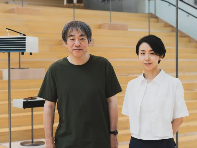 一名穿墨绿短袖的男子与一名穿白色短袖衬衫的女子并排站在一座浅色木阶梯前,阶梯上立着几台唱机