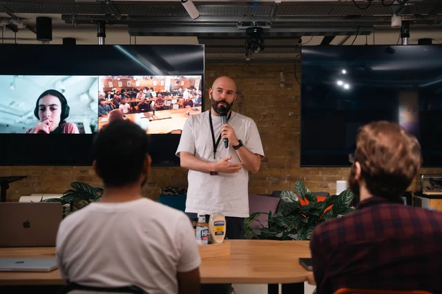 Speaker presenting at an evening tech conference with a screen and audience in the background