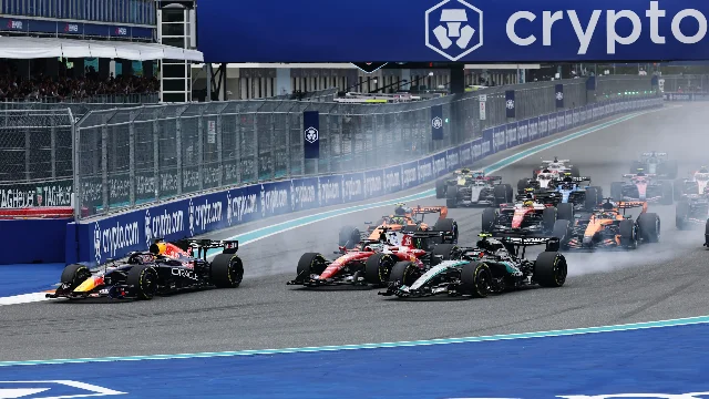Multiple F1 cars battling through Turn 1 at the Miami International Autodrome — Red Bull, Ferrari, Mercedes, and McLaren visible, grandstands packed
