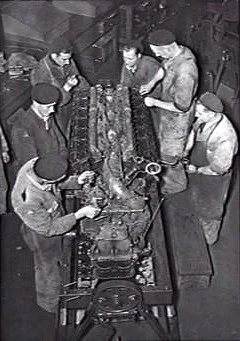 Six mechanics working around an aircraft engine in a wartime workshop