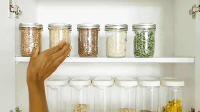 Glass jars of grains and clear containers of pulses on two shelves of a white cupboard, with a hand reaching toward the top row