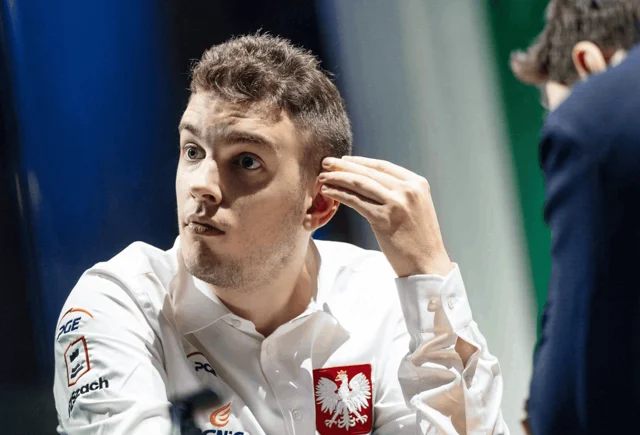 A chess player in a white shirt with a Polish crest on the sleeve, hand raised to his ear