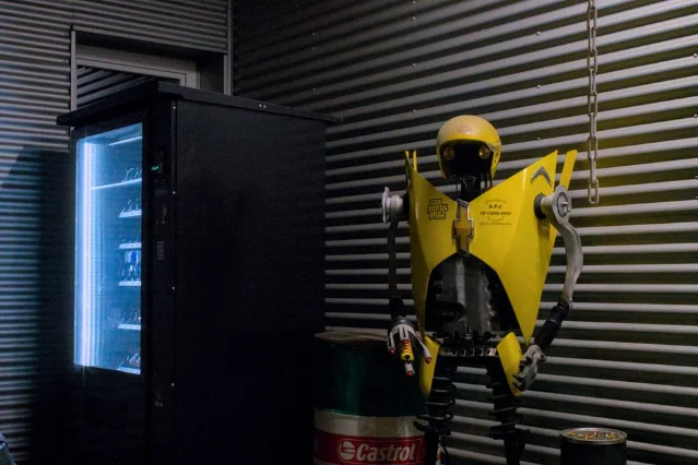A dimly lit industrial room with a vending machine beside a quirky robot sculpture, evoking the Andon Labs experimental setup
