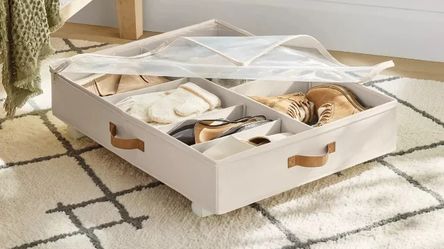 A cream fabric under-bed storage bin with its clear zippered top open, showing adjustable dividers holding shoes and folded knitwear, with casters visible on the base and the corner of a bed behind it.