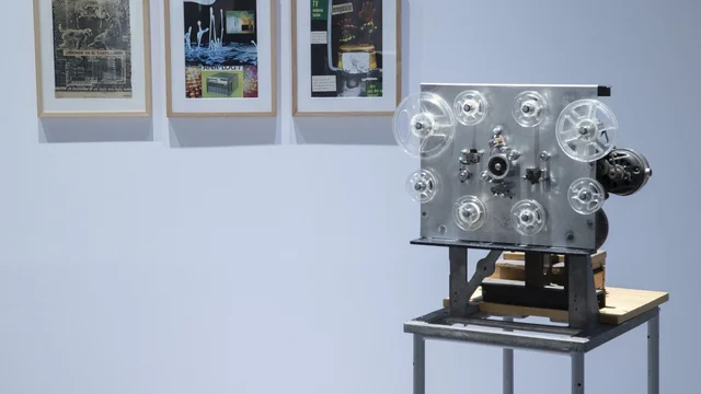 展厅里一台自制机械装置立在铁架上,几个透明片盘与齿轮外露,背景墙上挂着三幅印刷拼贴