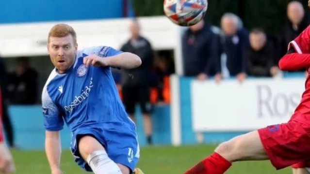 Martyn Jackson in a blue Ramsbottom United shirt challenging for the ball, with spectators behind the pitch rail