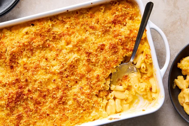 An overhead photograph of a white baking dish packed with macaroni and cheese under a deep golden crumb topping, a metal serving spoon lifting out a portion and revealing the loose cheese sauce and elbows beneath, with a second dark bowl of macaroni at the right edge