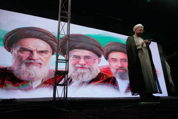 A cleric checks his cell phone on stage in front of a screen displaying portraits of late Supreme Leader Ayatollah Khamenei and Supreme Leader Ayatollah Mojtaba Khamenei during a pro-government gathering in Tehran, Iran, June 7, 2026.