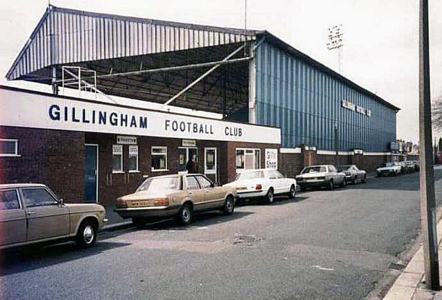 "The exterior of an old football ground: a stand with corrugated cladding above a brick frontage lettered GILLINGHAM FOOTBALL CLUB, with cars parked along the road"