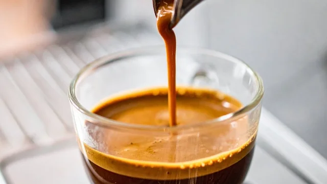 An espresso shot being poured from a portafilter into a clear glass cup, showing rich dark crema over a caramel-colored body