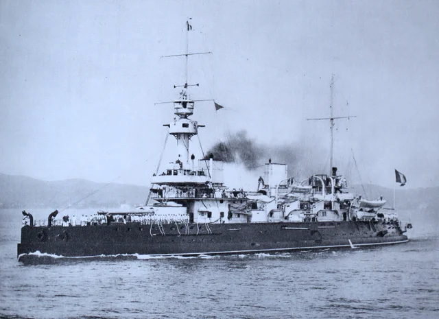 A tinted photograph of a warship moving at speed, her crew in white uniforms lined along the deck and a French flag at the stern