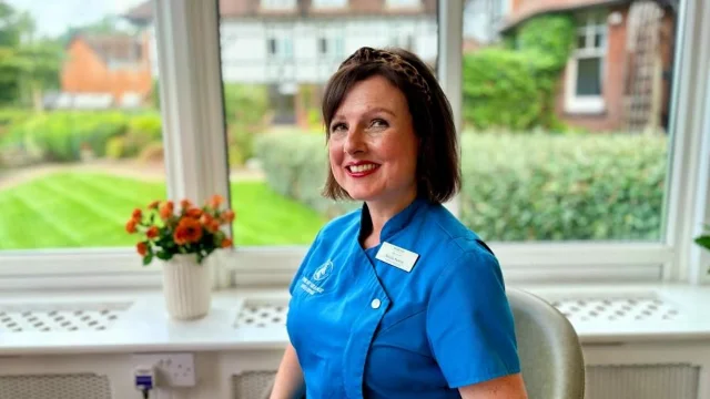 A woman in a blue care-home tunic smiling, seated by a window with orange flowers on the sill