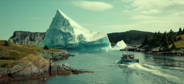 A fishing boat crossing calm water in front of a large iceberg, with rocky shoreline on either side, in Below