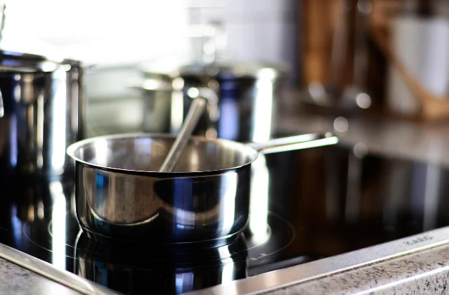 A stainless steel pan with a spoon in it sitting on a black glass hob, with other pans blurred behind