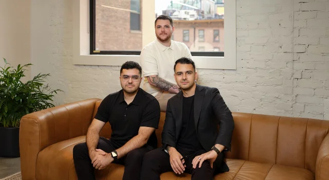 Three men seated and standing in a brick-walled office
