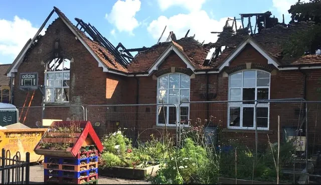 the burnt shell of a red brick school building, roof timbers exposed and windows gone, with fencing and a flower planter in front