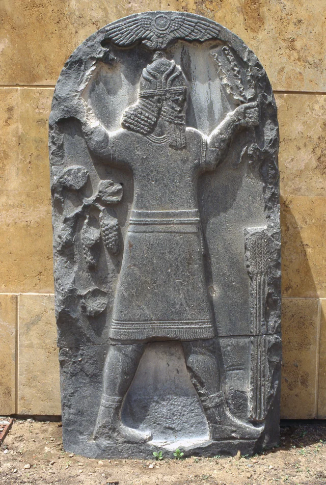 A dark stone stele carved with a standing figure holding grapes and ears of grain beneath a winged sun disc