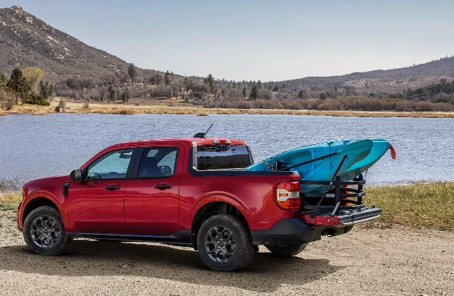 A red Ford Maverick pickup with two kayaks in the bed, parked by a lake below hills