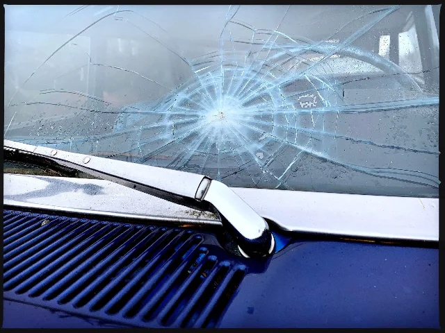 A windshield with a large impact point at the centre of a web of cracks radiating across most of the glass, above a chrome wiper arm and a dark blue car body