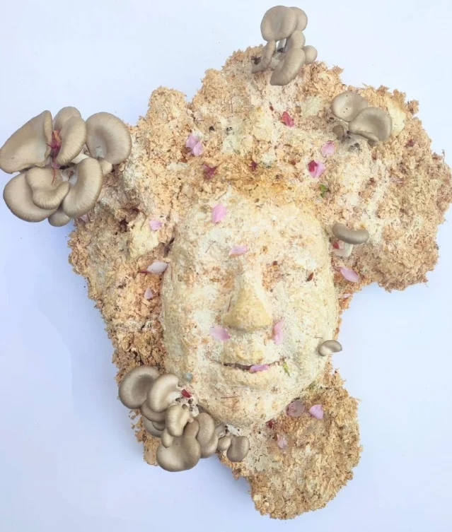 A pale face-shaped mycelium sculpture with clusters of mushrooms and scattered pink petals