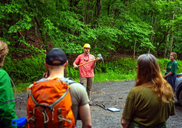 A trail-building instructor holds a Pulaski tool while volunteers listen in a green forest