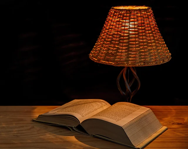 A table lamp with a woven shade beside an open book on a wooden surface