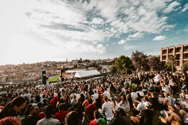 A large crowd gathers outdoors watching a soccer match on a big screen under a summer sky