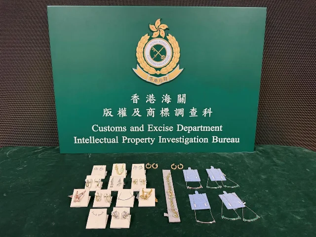 The Hong Kong Customs and Excise Department Intellectual Property Investigation Bureau display board on a green table, with 21 pieces of gold and silver jewellery laid out on white presentation cards in front of it
