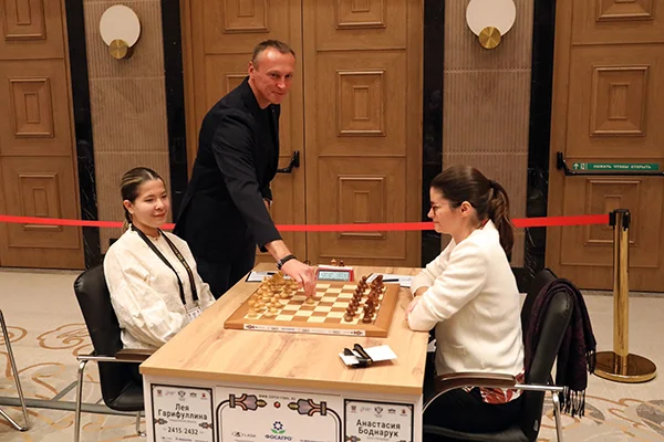 Two players seated at a board in a hotel hall with a man in a suit standing between them