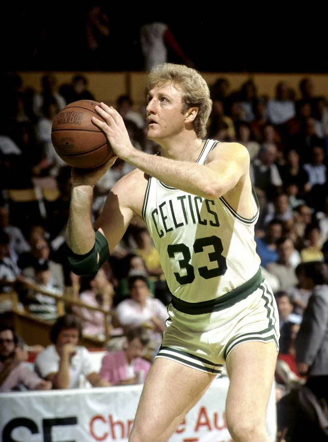 A basketball player in a white Celtics jersey looking up as he brings the ball up to shoot, with spectators behind him