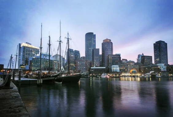 Boston waterfront at dusk — tall-masted ships docked at left, city skyline reflecting in calm harbor water