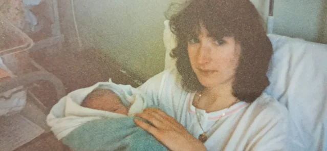 A young woman in a hospital bed holding a swaddled newborn against her chest, with a bassinet beside them.