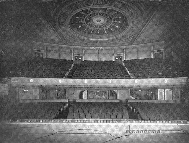 A grainy black-and-white photograph taken from a stage, looking out over two tiers of empty seats toward an ornate domed ceiling